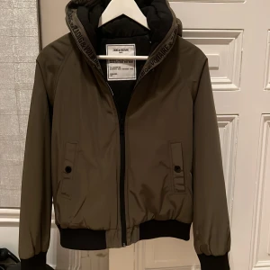 Olivgrön jacka från Zadig & Voltaire - Säljer en snygg olivgrön jacka från Zadig & Voltaire. Den har en dragkedja framtill och en huva med märkesdetaljer. Jackan är fodrad. Storlek 14 år
