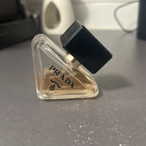 Parfym från Prada - Säljer en elegant parfym Prada paradoxe i en unik trekantig flaska. Den har en stilren design med en svart fyrkantig kork och en ljus doftvätska. Perfekt för den som vill ha en lyxig touch i sin doftsamling.