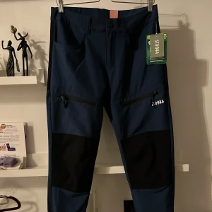 Blå byxor från Strøva - Säljer ett par snygga blå byxor från Strøva i nytt skick. De har svarta detaljer vid knäna och dragkedjor på fickorna. Perfekta för utomhusaktiviteter med en bekväm passform. Märkta med Bionic Finish Eco för extra hållbarhet.