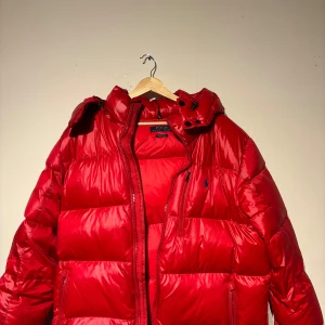 Röd vinterjacka från Polo Ralph Lauren - Säljer en snygg röd vinterjacka från Polo Ralph Lauren i storlek L. Jackan är perfekt för vintern med sin varma och puffiga design. Den har en dragkedja framtill och en justerbar huva. Märket är broderat på bröstet. Perfekt för kalla dagar! 🔥Skick 10/10