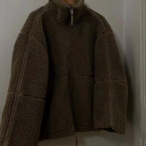 Brun teddyjacka med dragkedja - Mysig brun teddyjacka i mycket bra skick. Perfekt för höst och vinter med sin varma och fluffiga design. Jackan har en dragkedja framtill och långa ärmar, vilket gör den både praktisk och stilren. Perfekt för att hålla värmen under kyliga dagar!