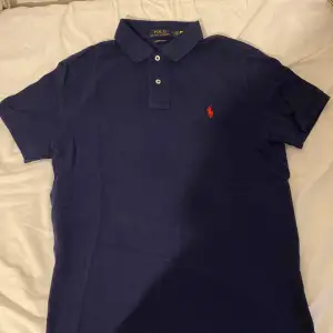 Säljer en snygg mörkblå pikétröja från Ralph Lauren i storlek M. Tröjan har en klassisk krage med två knappar och det ikoniska röda logotypbroderiet på bröstet. Perfekt för både vardag och fest! 💙