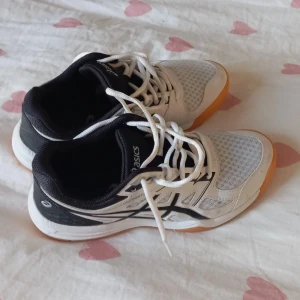 Vita och svarta Asics sneakers - Säljer ett par vita och svarta Asics sneakers i bra skick. De har en snygg design med snörning och en bekväm passform. Perfekta för träning eller vardagsbruk. Sulan är i en cool orange färg som ger extra stilpoäng.