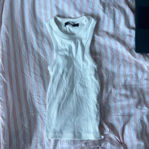 Vit ribbad linnetopp - Säljer en stilren vit ribbad linnetopp i nyskick. Perfekt för sommardagar eller som ett basplagg i garderoben. Toppen är ärmlös och har en tight passform som framhäver figuren. Superbekväm och lätt att matcha med olika outfits!