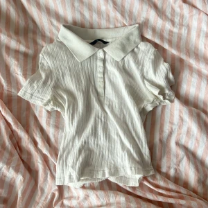 Vit ribbad polotopp - Säljer en söt vit ribbad polotopp med korta ärmar och knappar framtill. Perfekt för vår och sommar, och funkar både till vardags och lite finare tillfällen. Toppen är i bra skick och superbekväm att ha på sig. Säljer pga att den inte kommer till användning <3