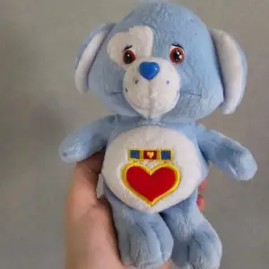 En blå Care Bear-plyschleksak med ett rött hjärta på bröstet. Björnen (hunden) är ca 20 cm hög och i gott skick. Den är från 2003.   Sökord Samlarobjekt  Care Bear  Vintage Gosedjur 