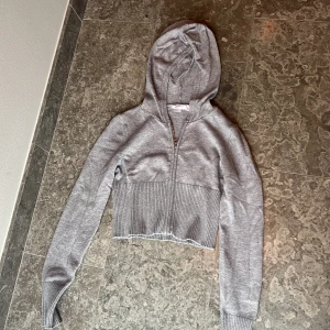 Grå croppad hoodie från Gina Tricot - Säljer en superfin grå croppad hoodie från Gina Tricot. Den har en dragkedja framtill och en mysig huva. Perfekt för en avslappnad stil och passar bra till både jeans och kjol. Den ribbade nederkanten ger en snygg detalj. Perfekt för höst och vår!