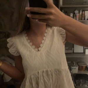 Beige blus med spetsdetaljer - Säljer en söt beige blus med v-ringning och spetsdetaljer längs halsringningen. Blusen har korta ärmar med volang och ett lätt, luftigt material som passar perfekt för sommardagar. Den har en lös passform som gör den bekväm och stilren.