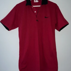 Röd pikétröja från Nike - Säljer en snygg röd pikétröja från Nike. Den har en klassisk krage i svart med en knapp och det ikoniska Nike-loggan broderad på bröstet. Perfekt för både vardag och sportiga aktiviteter. Passar bra till jeans eller shorts! Den är ganska nopprig men med lite tålamod man man nog få den fin igen.