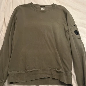 Olivgrön tröja från C.P. Company - Säljer en olivgrön tröja från C.P. Company i mycket bra skick. Den har en cool fickdetalj med knapp på ärmen och är perfekt för höst och vår. Tröjan är långärmad och har ribbade muddar vid ärmslut och nederkant. Perfekt för en avslappnad stil!