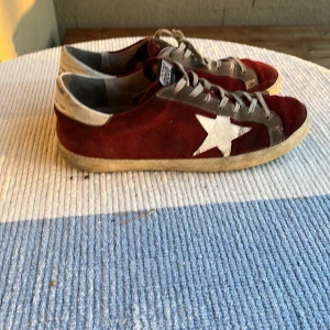 Golfen goose vinröd - Säljer ett par coola röda sneakers från golden goose med en vit stjärna på sidan. Skorna har grå snörning och en sliten look för en avslappnad stil. Perfekta för både vardag och fest!