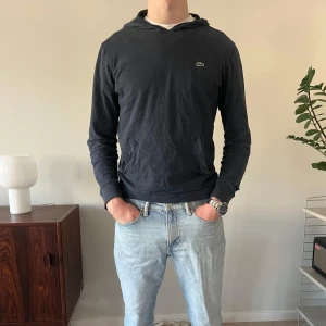 Grå-blå Lacoste-Hoodie  - Mycket bekväm och snygg hoddie från Lacoste.  Mycket fint skick. Två små deffekter i form av två mycket små hål vid tröjans ”midjefickor”. (Se bilderna) Dessa syns dock inte i allmänhet när man bär tröjan likt på bilderna. 