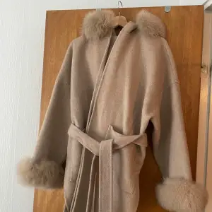 Säljer en superfin beige kappa med fluffiga pälsdetaljer på krage och ärmslut. Den har ett bälte i midjan för en snygg passform. Perfekt för höst och vinter när du vill vara både varm och stilren. Passar till alla tillfällen!