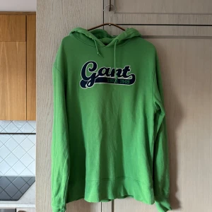 Grön hoodie från Gant - Storlek L - Säljer en snygg grön hoodie från Gant med stort tryck på framsidan. Den är i  bra använt skick och perfekt för både höst och vår. Hoodien har en klassisk passform med långa ärmar och en skön huva med dragsko. Perfekt för en avslappnad stil!