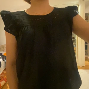 Svart blus med spetsdetaljer - Säljer en svart blus med vackra spetsdetaljer vid ärmarna. Blusen har en luftig passform och korta ärmar med volang. Perfekt för både vardag och festliga tillfällen.