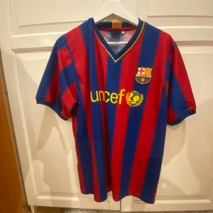 Blåröd FC Barcelona fotbollströja med Ibrahimović 9 - Säljer en klassisk FC Barcelona fotbollströja i blårött med gula detaljer. Tröjan har korta ärmar och ett randigt mönster. På framsidan finns klubbmärket och Unicef-loggan, och på baksidan står Ibrahimović 9. 