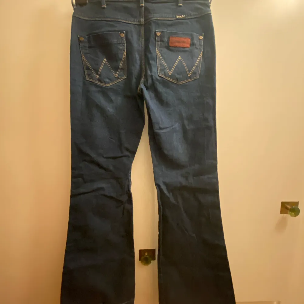 Säljer ett par klassiska mörkblå/denim bootcut jeans från Wrangler. Snygg passform med mid/highrise. Fina detaljer. Är 163cm lång och byxorna går ner i golvet, har därför vikt upp dem vilket resulterat i lite slitage (som knappt syns, ljusare streck där byxorna varit vikta). Riktigt coola 70-tals jeans! ⭐️⭐️💞. Farkut & Housut.