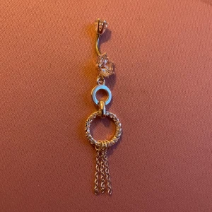 Navelpersing med kristall och kedjor - Elegant silver navelpersing med en stor klar kristall och en cirkel dekorerad med små gnistrande stenar. Från cirkeln hänger tre tunna kedjor som ger en fin rörelse. Perfekt för att lägga till lite glamour till din look.