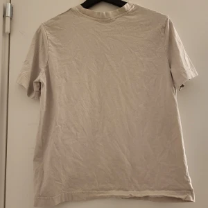Beige t-shirt med tryck - Säljer en beige t-shirt med texten 'Gala de la Palmera' i svart tryck på framsidan. T-shirten har en klassisk rund hals och korta ärmar. Perfekt för en avslappnad stil.
