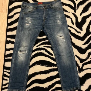 Blå jeans med slitningar - Snygga blå jeans med coola slitningar och en klassisk femficksdesign. Perfekta grischbyxor. De har en knappgylf och är tillverkade i ett robust denimtyg🥵