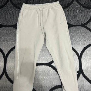 Beige mjukisbyxor från Boss - Säljer ett par stilrena beige mjukisbyxor från Boss. Byxorna har en elastisk midja med dragsko och diskret logga på sidan. Perfekta för en avslappnad look om ni köper hela tracksuit kmr de kåsta bara 699