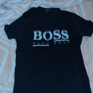 Svart t-shirt från Hugo Boss - Snygg svart t-shirt från Hugo Boss med stort BOSS-tryck på framsidan. Perfekt för en stilren look. Jätte liten Tshirt 