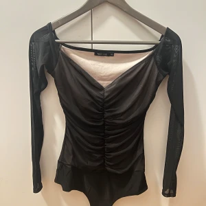 Svart mesh body topp från NLY One - Snygg svart mesh body topp från NLY One i storlek XS. Toppen har långa ärmar och en v-ringad design med rynkdetaljer framtill. Perfekt för en kväll ute eller en speciell tillställning.