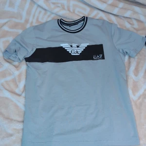 Grå t-shirt från EA7 H - Snygg grå t-shirt från EA7 med svart och vitt logotryck på bröstet. Den har en rund halsringning med svarta och vita detaljer. Perfekt för en avslappnad stil.