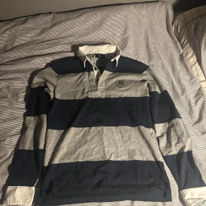 Randig pikétröja från GANT - Säljer en stilren randig pikétröja från GANT i marinblått och grått. Tröjan har långa ärmar och en klassisk krage med knappar. Perfekt för en avslappnad men ändå snygg look.