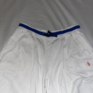 Vita shorts från Polo Ralph Lauren - Säljer ett par vita shorts från Polo Ralph Lauren med blå detaljer och snörning i midjan. Shortsen har fickor fram och en broderad logga i orange. Perfekta för sommardagar!