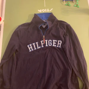 Svart tröja med dragkedja från Tommy Hilfiger - Säljer en svart långärmad tröja från Tommy Hilfiger med dragkedja och blå detaljer. Tröjan har ett stort Hilfiger-tryck på framsidan och en amerikansk flagga på ärmen. Perfekt för en avslappnad stil.