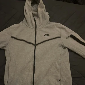 Grå nike tech fleece köpt från 1200kr har ett litet hål som man knappt ser och den är lite syd annars bra skick 7/10 snabb affär  - Säljer en grå huvtröja från Nike med dragkedja och svart logotyp på bröstet. Tröjan har långa ärmar och en praktisk huva. Perfekt för en avslappnad stil.