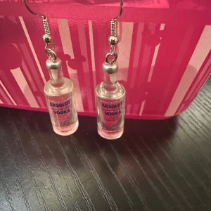 Örhängen med Absolut Vodka-flaskor - Unika örhängen formade som små Absolut Vodka-flaskor. Perfekta för den som vill ha en rolig och annorlunda accessoar. Flaskorna är detaljerade och hänger från silverfärgade krokar.