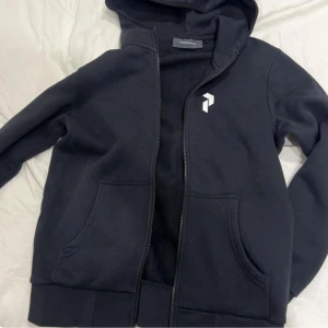 Svart huvtröja med dragkedja - Säljer en svart huvtröja från Peak Performance med dragkedja och en liten logga på bröstet. Tröjan har långa ärmar och en praktisk huva. Perfekt för en avslappnad stil. STORLEK: S