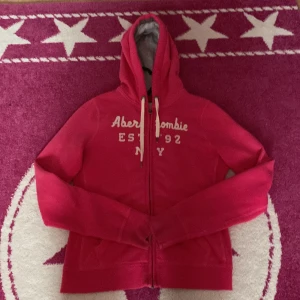 Rosa hoodie från Abercrombie & Fitch - Säljer en snygg rosa hoodie från Abercrombie & Fitch med vit text på framsidan. Hoodien har en dragkedja och justerbara snören i huvan. Perfekt för Y2K, Mcbling, Drainer och annat poppis stil