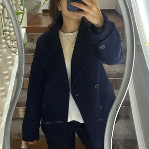 Mörkblå kappa från Nelly - Snygg mörkblå kappa från Nelly med dubbelknäppning och breda slag. Perfekt för kyligare dagar med sin stilrena design och långa ärmar. Ett måste i garderoben för en klassisk look.