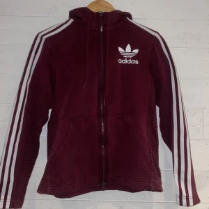 Vinröd hoodie från Adidas - Säljer en vinröd hoodie från Adidas med klassiska vita ränder på ärmarna och logga på bröstet. Hoodien har dragkedja och en praktisk huva med snörning. Perfekt för en sportig look. XS men passar definitivt S.
