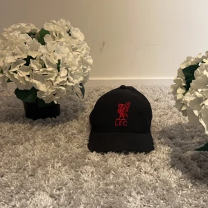Svart keps med röd LFC-logga - Snygg svart keps med röd broderad LFC-logga framtill. Märket 47 syns på sidan. Perfekt för fotbollsfans som vill visa sitt stöd med stil. Pris kan diskuteras