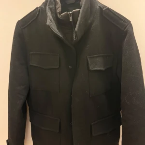 Svart jacka från Jack & Jones - Snygg svart jacka från Jack & Jones med quiltad insida och praktiska fickor. Jackan har en dragkedja och knappar framtill samt axelklaffar för en stilren look. Perfekt för kyligare dagar.