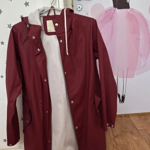 Vinröd regnjacka från 157 - Snygg vinröd regnjacka från 157 med huva och knappar. Perfekt för regniga dagar med sin långa design och praktiska fickor. Jackan har en stilren look och är lätt att matcha med olika outfits.