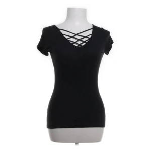 Svart topp med korsdetalj från Hollister - Snygg svart topp från Hollister med korsdetalj vid v-ringningen. Perfekt för en stilren look. Kortärmad och i en bekväm passform.