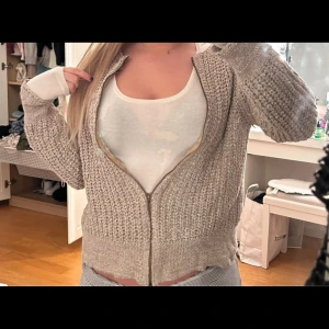 Beige aktig stickad kofta - Mysig beige stickad kofta med dragkedja framtill. Koftan har långa ärmar och en ribbad struktur som ger en skön och avslappnad passform. Den är lite guld aktig kofta 