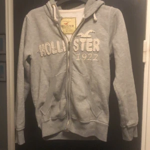 Grå hoodie från Hollister - Säljer en grå hoodie från Hollister med dragkedja och broderad logga på framsidan. Tröjan har långa ärmar och en mysig huva med snören. Perfekt för en avslappnad stil.