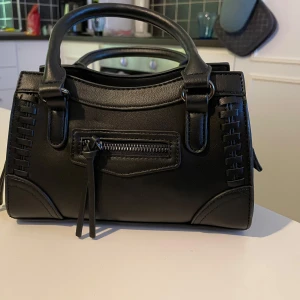 Svart handväska med dragkedja - Snygg svart handväska i syntetmaterial med praktisk dragkedja och dekorativa detaljer. Perfekt för att bära dina dagliga nödvändigheter med stil. Helt oanvänd nyskick