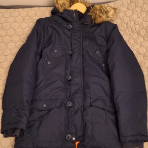 Mörkblå dunjacka från Polo Ralph Lauren - Säljer en mörkblå dunjacka från Polo Ralph Lauren med fuskpäls på huvan. Jackan har knappar och dragkedja framtill samt flera praktiska fickor. Perfekt för kalla vinterdagar.