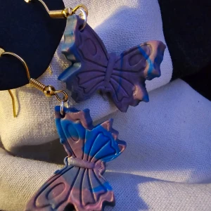 Fjärilsformade örhängen i lila och blått - Unika fjärilsformade örhängen i en mix av lila och blå nyanser. De har en kreativ design med en marmorerad effekt och är fästa på guldfärgade krokar. Perfekta för att lägga till en färgglad touch till din stil. Guldpläterade.