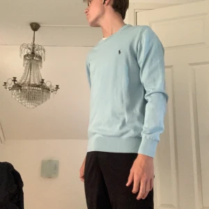 Ljusblå tröja från Ralph Lauren - Säljer en stilren ljusblå tröja från Ralph Lauren med rund hals och det klassiska logotypbroderiet på bröstet. Tröjan har långa ärmar och ribbade muddar vid ärmslut och nederkant. Perfekt för en avslappnad men ändå elegant look.