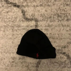 Svart mössa från Ralph Lauren - Snygg svart mössa från Ralph Lauren med röd broderad logga framtill. Perfekt för kyliga dagar och en stilren look.
