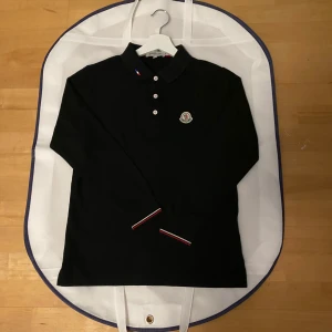 Svart pikétröja från Moncler - Snygg svart pikétröja från Moncler med klassisk krage och tre knappar. Tröjan har Monclers logotyp på bröstet och detaljer i rött, vitt och blått vid kragen och ärmsluten. Perfekt för en stilren look. Passar 165-175