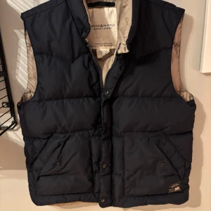 Ralph Lauren Denim & Supply väst - Säljer en snygg mörkblå dunväst från Ralph Lauren, Denim & Supply. Västen har en dragkedja och knappar framtill samt två praktiska fickor. Perfekt för kyliga dagar.